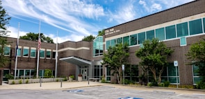 The image depicts a modern brick building identified as a city hall. It features a symmetrical facade with numerous windows and a prominent entrance. The surrounding area includes a well-maintained landscape with small trees and shrubs, along with accessible parking spaces. Three flagpoles are positioned in front of the building, displaying the flags.