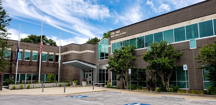 The image depicts a modern brick building identified as a city hall. It features a symmetrical facade with numerous windows and a prominent entrance. The surrounding area includes a well-maintained landscape with small trees and shrubs, along with accessible parking spaces. Three flagpoles are positioned in front of the building, displaying the flags.