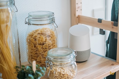 Jars filled with pasta and grains are placed on a wooden shelf next to a small plant. A white smart speaker sits alongside. The shelf is part of a light-colored wooden storage unit, creating a cozy kitchen atmosphere.
