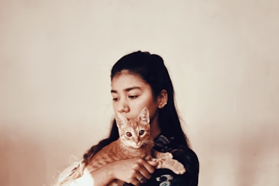 A young woman with long dark hair holds an orange tabby cat close to her chest. The cat looks towards the camera with wide eyes, while the woman gazes off to the side in a contemplative manner. The background is a soft, neutral color, adding a warm, intimate atmosphere to the scene.