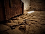 An old, rustic lock and two keys are lying on a stone floor. A wooden chest with visible aging and texture is situated nearby. The lighting is dim with a narrow beam of light illuminating part of the floor.