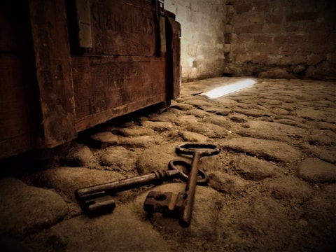 An old, rustic lock and two keys are lying on a stone floor. A wooden chest with visible aging and texture is situated nearby. The lighting is dim with a narrow beam of light illuminating part of the floor.