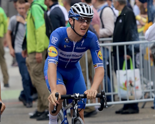 A cyclist wearing a blue uniform and helmet is riding a bicycle. The cyclist appears focused and is surrounded by spectators and other individuals in the background. Barriers are seen along the side of the street suggesting an organized event or race.