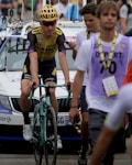 A cyclist wearing a yellow and black racing outfit with a matching helmet is riding a Bianchi bike, followed by people who appear to be in a race setting. Behind them, a white support vehicle with a rooftop rack is visible, and a person wearing a purple vest marked 'DEPART 70' walks nearby.