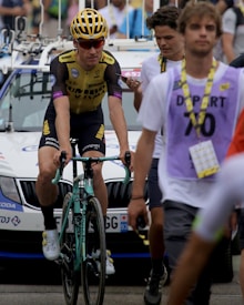 A cyclist wearing a yellow and black racing outfit with a matching helmet is riding a Bianchi bike, followed by people who appear to be in a race setting. Behind them, a white support vehicle with a rooftop rack is visible, and a person wearing a purple vest marked 'DEPART 70' walks nearby.