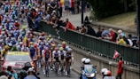 A large group of cyclists in a professional race surrounded by a crowd of spectators. The cyclists are wearing colorful jerseys and helmets, indicating teams or sponsors. A support vehicle and a motorcycle with officials or security are also present, indicating this is part of an organized event.