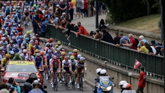 A large group of cyclists in a professional race surrounded by a crowd of spectators. The cyclists are wearing colorful jerseys and helmets, indicating teams or sponsors. A support vehicle and a motorcycle with officials or security are also present, indicating this is part of an organized event.