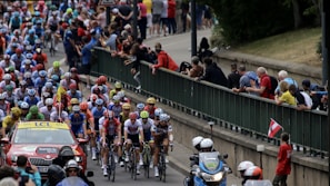 A large group of cyclists in a professional race surrounded by a crowd of spectators. The cyclists are wearing colorful jerseys and helmets, indicating teams or sponsors. A support vehicle and a motorcycle with officials or security are also present, indicating this is part of an organized event.