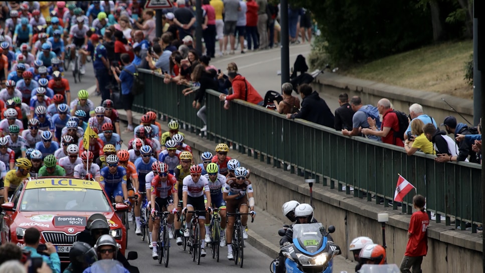 A large group of cyclists in a professional race surrounded by a crowd of spectators. The cyclists are wearing colorful jerseys and helmets, indicating teams or sponsors. A support vehicle and a motorcycle with officials or security are also present, indicating this is part of an organized event.