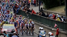A large group of cyclists in a professional race surrounded by a crowd of spectators. The cyclists are wearing colorful jerseys and helmets, indicating teams or sponsors. A support vehicle and a motorcycle with officials or security are also present, indicating this is part of an organized event.
