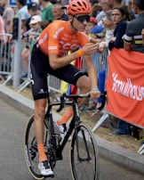 A cyclist in an orange and black outfit rides a road bicycle during a competitive cycling event. Spectators are gathered along the sidelines, some leaning over barriers to watch or cheer. The cyclist wears an orange helmet and sunglasses, with visible brands on the jersey and bike.