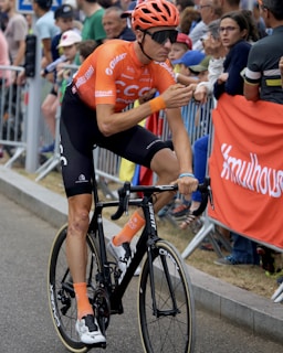 A cyclist in an orange and black outfit rides a road bicycle during a competitive cycling event. Spectators are gathered along the sidelines, some leaning over barriers to watch or cheer. The cyclist wears an orange helmet and sunglasses, with visible brands on the jersey and bike.