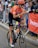 A cyclist in an orange and black outfit rides a road bicycle during a competitive cycling event. Spectators are gathered along the sidelines, some leaning over barriers to watch or cheer. The cyclist wears an orange helmet and sunglasses, with visible brands on the jersey and bike.