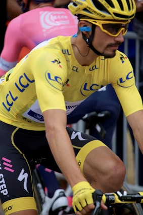 A cyclist wearing a bright yellow jersey and matching helmet is seen in motion, participating in a race. The jersey displays various logos and lettering in blue. The individual has on yellow gloves and sunglasses, with a focused and determined expression. Another cyclist in a pink attire is visible in the background, indicating a competitive environment.