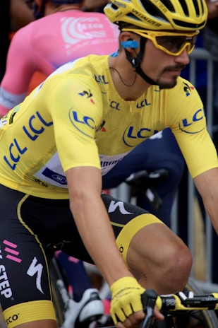 A cyclist wearing a bright yellow jersey and matching helmet is seen in motion, participating in a race. The jersey displays various logos and lettering in blue. The individual has on yellow gloves and sunglasses, with a focused and determined expression. Another cyclist in a pink attire is visible in the background, indicating a competitive environment.