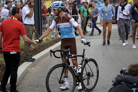 A cyclist wearing a race uniform and helmet shakes hands with another person in a red shirt. The cyclist is holding a black racing bike. The scene is outdoors, and there are several people in the background, some appearing to be spectators and officials. The atmosphere suggests a cycling event or race.