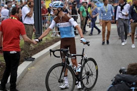 A cyclist wearing a race uniform and helmet shakes hands with another person in a red shirt. The cyclist is holding a black racing bike. The scene is outdoors, and there are several people in the background, some appearing to be spectators and officials. The atmosphere suggests a cycling event or race.