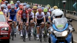 A group of cyclists tightly packed together are participating in a race. They are wearing colorful jerseys and helmets, indicating a competitive cycling event. A motorbike and a support car with signage accompany them on a road that runs alongside a railing or barrier.