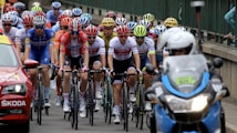 A group of cyclists tightly packed together are participating in a race. They are wearing colorful jerseys and helmets, indicating a competitive cycling event. A motorbike and a support car with signage accompany them on a road that runs alongside a railing or barrier.