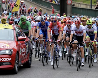 A large group of cyclists is participating in a race on a city street, closely followed by a red support car. The cyclists wear colorful jerseys and helmets, creating a vibrant scene. The atmosphere is dynamic and competitive.
