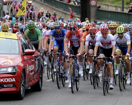 A large group of cyclists is participating in a race on a city street, closely followed by a red support car. The cyclists wear colorful jerseys and helmets, creating a vibrant scene. The atmosphere is dynamic and competitive.