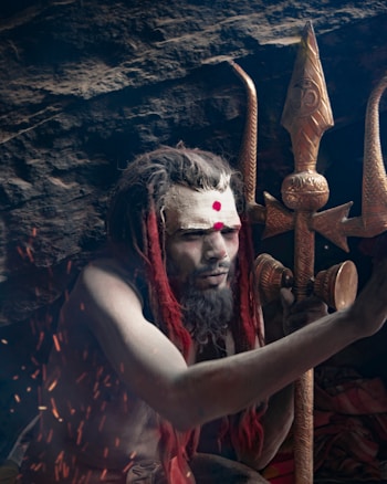 A person with dreadlocked hair and ash-painted face, adorned with red markings, is sitting in a meditative posture. The individual is holding a trident, which is elaborately designed with intricate carvings. The background consists of a rocky surface, with sparks of fire visible, adding to the mystical ambiance.