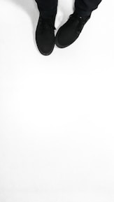 A pair of black shoes is worn by a person standing on a solid white surface. The shoes appear to be casual and are accompanied by black pants.