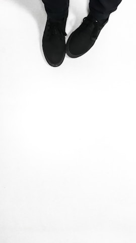 A pair of black shoes is worn by a person standing on a solid white surface. The shoes appear to be casual and are accompanied by black pants.