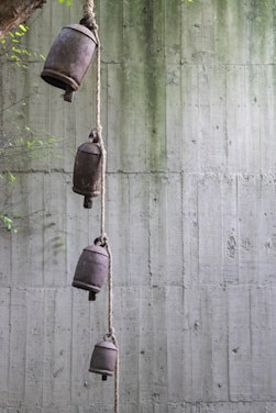 A series of four rustic bells tied vertically along a thick rope are hanging from a tree branch against a textured concrete wall. The bells have a weathered and aged appearance, contributing to an overall rustic feel. The background is a bare concrete wall with a subtle gradient of green from top to bottom, with some foliage visible on the left side of the image.