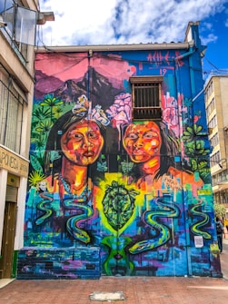 A vibrant street mural featuring two large, colorful portraits of women surrounded by an array of flowers and abstract shapes. The background includes lush green foliage, a mountain with pink hues, and a bright blue sky. The mural is painted on the side of a building, contributing to an urban artistic atmosphere.