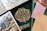 An assortment of books and papers featuring ornate designs and illustrations. The focal point is a green book titled 'The Book of Wisdom' with an intricate floral pattern. Surrounding items include a map, a drawing of a medieval landscape, a patterned envelope, and a card with printed text. The composition suggests themes of exploration, knowledge, and creativity.