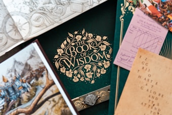 An assortment of books and papers featuring ornate designs and illustrations. The focal point is a green book titled 'The Book of Wisdom' with an intricate floral pattern. Surrounding items include a map, a drawing of a medieval landscape, a patterned envelope, and a card with printed text. The composition suggests themes of exploration, knowledge, and creativity.