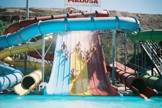 A group of people are sliding down large water slides of various colors into a pool at an outdoor water park. The slides are vibrant, and water splashes in the air as they descend. The backdrop includes a hilly terrain and clear blue sky.