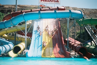 A group of people are sliding down large water slides of various colors into a pool at an outdoor water park. The slides are vibrant, and water splashes in the air as they descend. The backdrop includes a hilly terrain and clear blue sky.