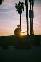 A silhouette of a person playing a guitar against a backdrop of a sunset. The sun is partially visible through the guitar, creating a warm glow. Tall palm trees and a street sign are in the background, adding to the serene atmosphere.