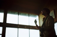 A person silhouetted against large windows holds a guitar, creating a moody and artistic atmosphere. The background is softly lit, highlighting the shape of the musician and the instrument.