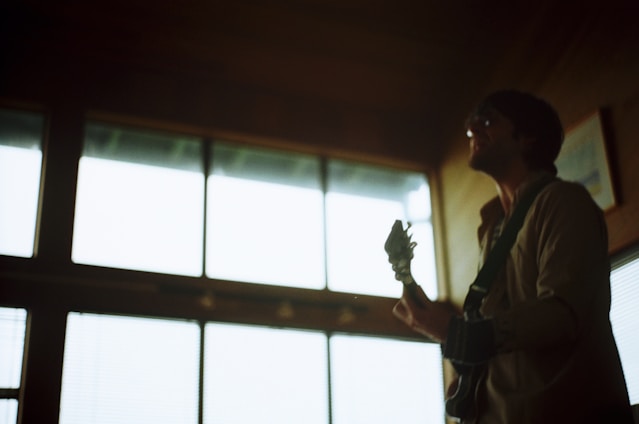 A person silhouetted against large windows holds a guitar, creating a moody and artistic atmosphere. The background is softly lit, highlighting the shape of the musician and the instrument.