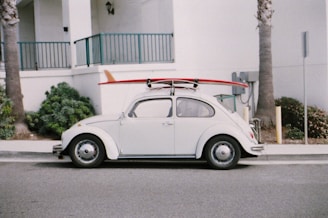 A vintage white Volkswagen Beetle is parked on the street with a red surfboard strapped to its roof. The car is positioned against a backdrop of a white building with teal railings, and there are palm trees nearby.