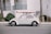 A vintage white Volkswagen Beetle is parked on the street with a red surfboard strapped to its roof. The car is positioned against a backdrop of a white building with teal railings, and there are palm trees nearby.