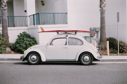A vintage white Volkswagen Beetle is parked on the street with a red surfboard strapped to its roof. The car is positioned against a backdrop of a white building with teal railings, and there are palm trees nearby.