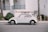 A vintage white Volkswagen Beetle is parked on the street with a red surfboard strapped to its roof. The car is positioned against a backdrop of a white building with teal railings, and there are palm trees nearby.