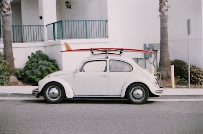 A vintage white Volkswagen Beetle is parked on the street with a red surfboard strapped to its roof. The car is positioned against a backdrop of a white building with teal railings, and there are palm trees nearby.