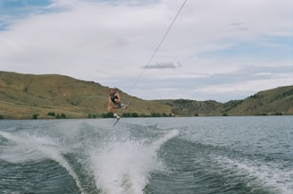 A person is wakeboarding on a large body of water, performing a jump with the wakeboard held firmly below their feet. The water is creating waves and splashes behind them, indicating motion and speed. Surrounding the lake are vast, hilly landscapes under a cloudy sky, adding to the scenic background.