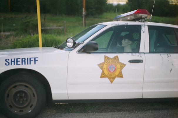 A police car labeled 'Sheriff' is parked on a roadside, displaying the Lake County Sheriff's badge on its side. Inside the vehicle, a mannequin wearing a cap is positioned in the driver's seat. The car shows signs of wear with chipped paint.