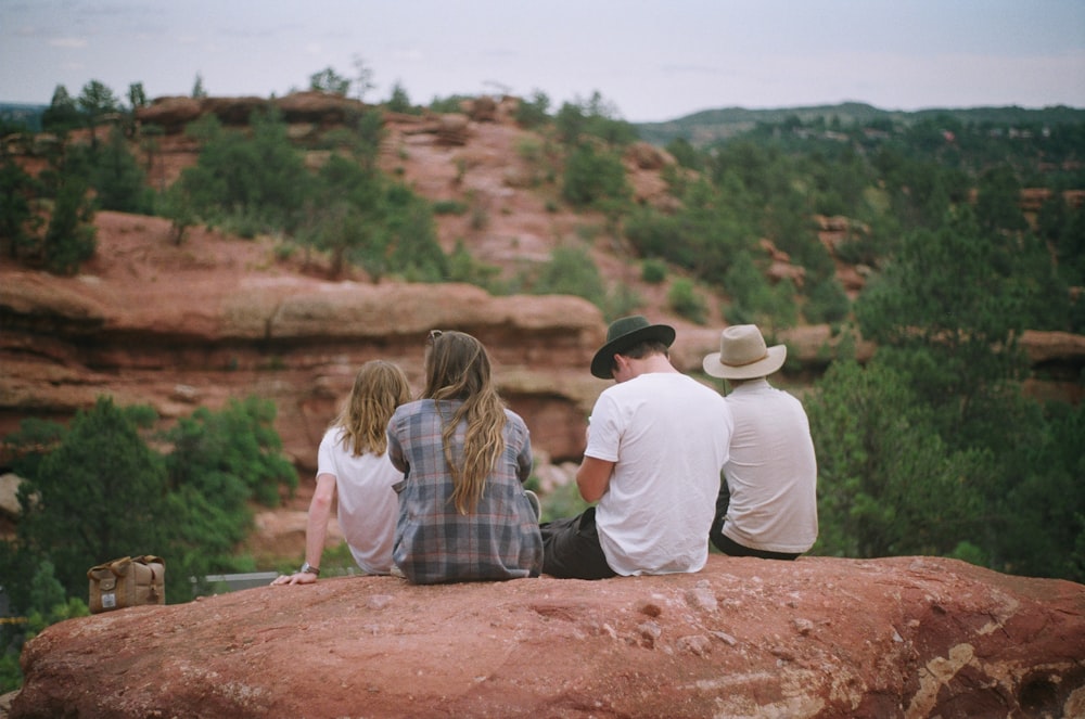 four person sitting on rock and facing hill with trees during daytime