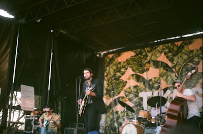 A band performs on an outdoor stage with a colorful, tropical backdrop. The lead singer plays a guitar while standing in front of a microphone. Another musician is seated playing an electric guitar, and a third musician is performing with a double bass. The stage is covered, and various musical equipment is visible.