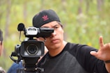 A person is operating a professional video camera, wearing a black cap with a pink logo and a black and gray shirt. The background is blurred, consisting of green trees or foliage, suggesting an outdoor setting. The individual appears focused on framing or directing a shot.
