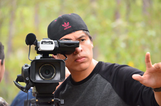 A person is operating a professional video camera, wearing a black cap with a pink logo and a black and gray shirt. The background is blurred, consisting of green trees or foliage, suggesting an outdoor setting. The individual appears focused on framing or directing a shot.