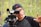 A person is operating a professional video camera, wearing a black cap with a pink logo and a black and gray shirt. The background is blurred, consisting of green trees or foliage, suggesting an outdoor setting. The individual appears focused on framing or directing a shot.