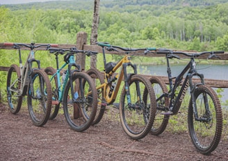 Four mountain bikes are lined up in a row on a dirt path next to a rustic, wooden fence. The background features a lush, green forest with a glimpse of a serene body of water, suggesting a natural and peaceful outdoor setting.